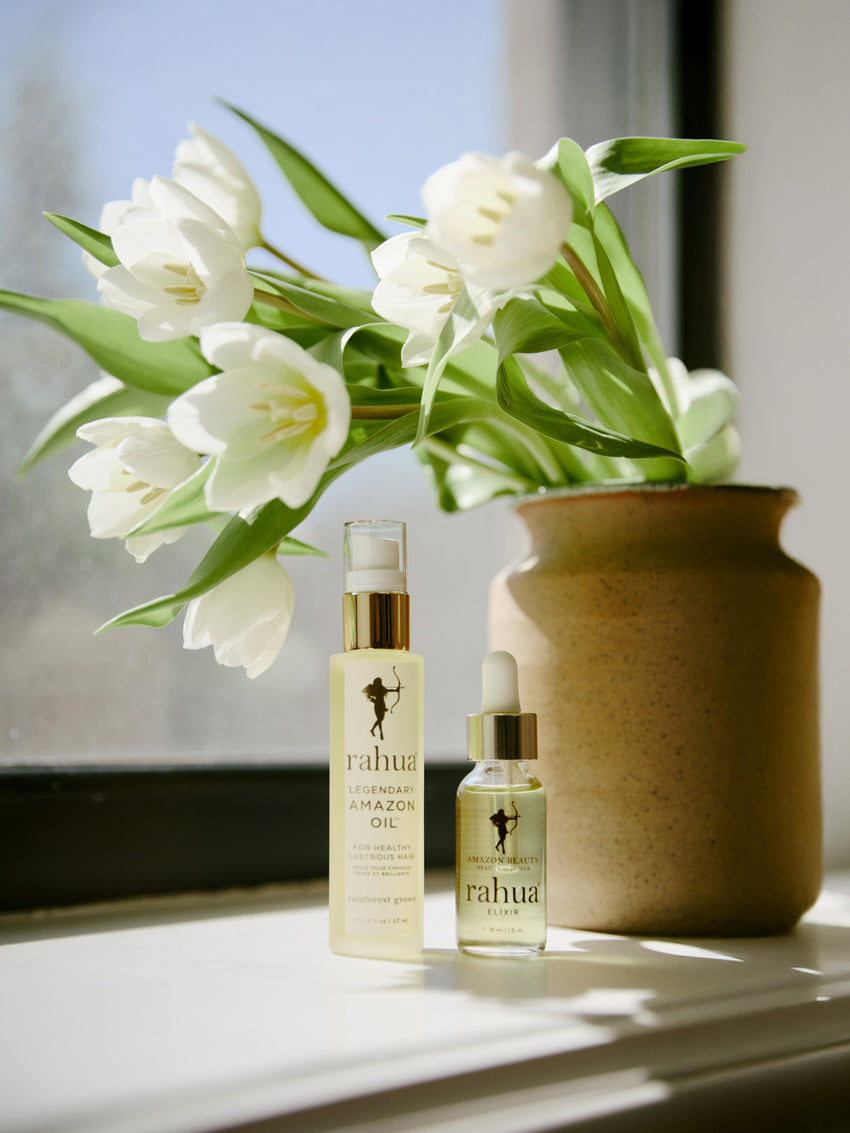 bottle of golden liquid in a glass bottle rahua elixir oil made of 100% rahua oil with a bottle of rahua legendary amazon oil on a window sill with a vase of tulips|variant:1-oz