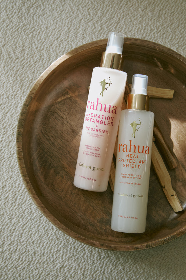 two bottles of a rahua hydration detangler and heat protectant shield in a wooden bowl with sticks of palo santo incense on a off white fabric background|variant:65-oz