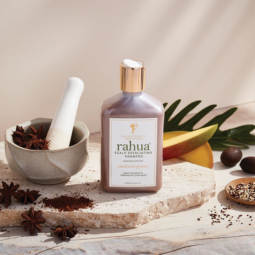 bottle of rahua scalp exfoliating shampoo sitting on a stone slab with a plain beige background with mangoes, quinoa, and star anise in a mortar and pestle being crushed|variant:93-oz