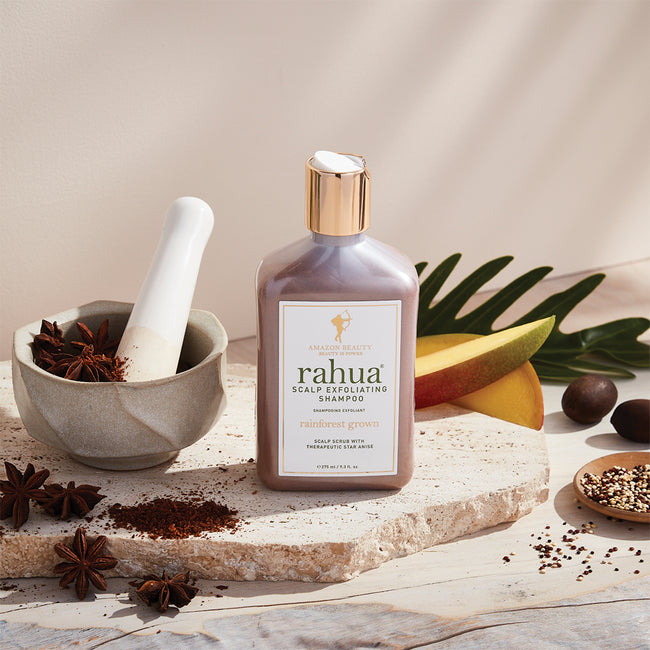 bottle of rahua scalp exfoliating shampoo sitting on a stone slab with a plain beige background with mangoes, quinoa, and star anise in a mortar and pestle being crushed|variant:93-oz