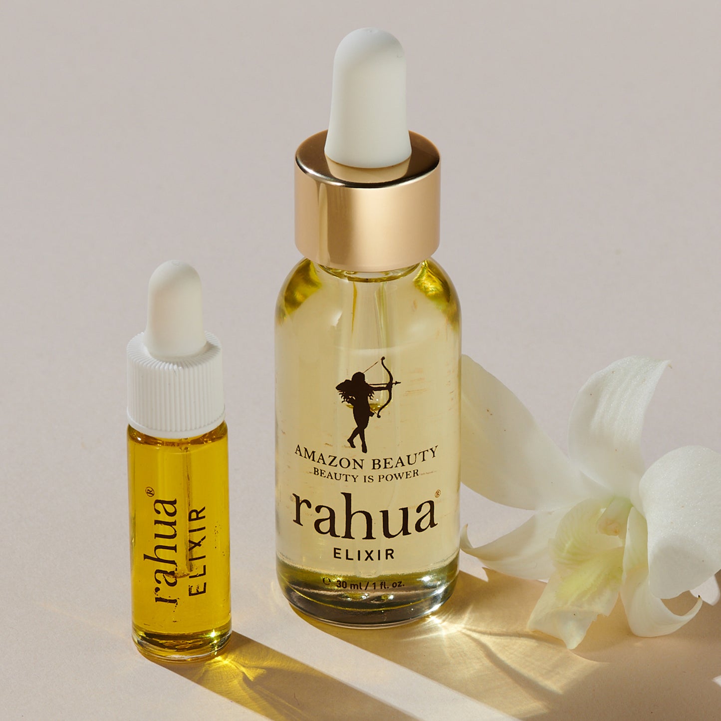 bottle of golden liquid in a glass bottle rahua elixir oil made of 100% rahua oil on a beige background with its pillow pack with gold rahua logo and elixir in brown|variant:015-oz