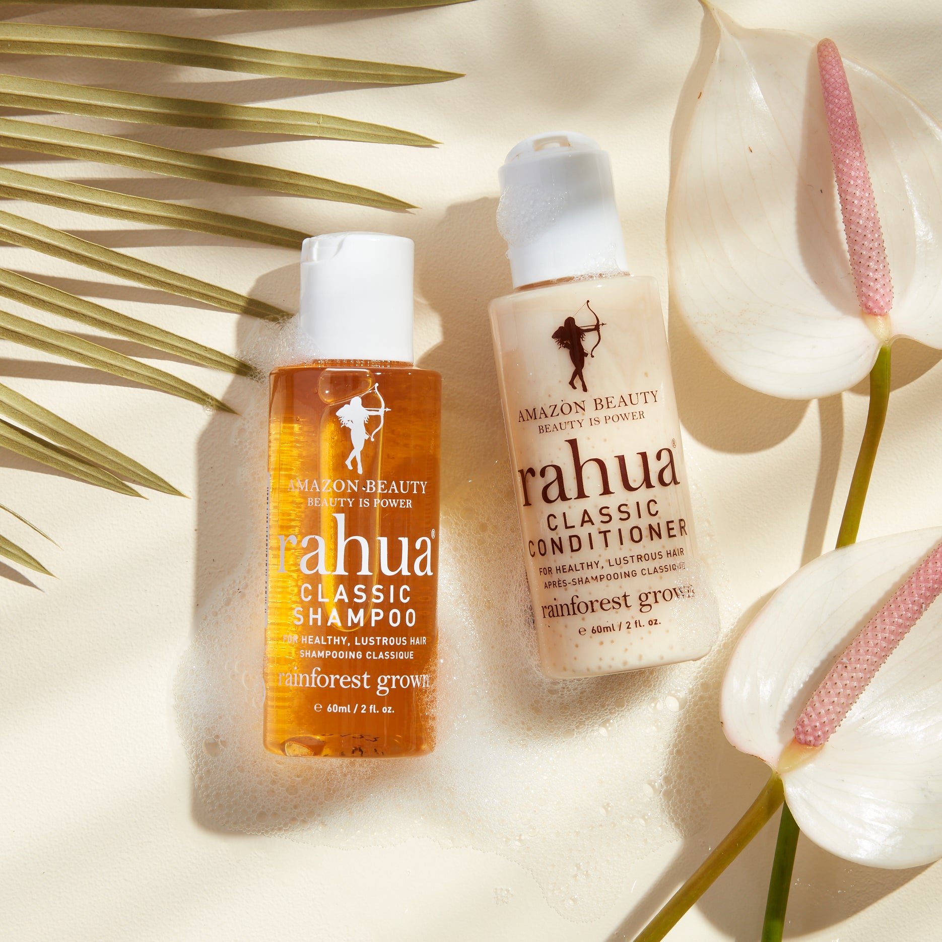 two plastic bottles of 2 oz rahua classic shampoo and classic conditioner with white caps on a soapy beige background and a palm travel friendly tsa safe|variant:2-oz